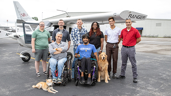 A group of people pose in front of an airplane. Two are in wheelchairs and each has a dog.
