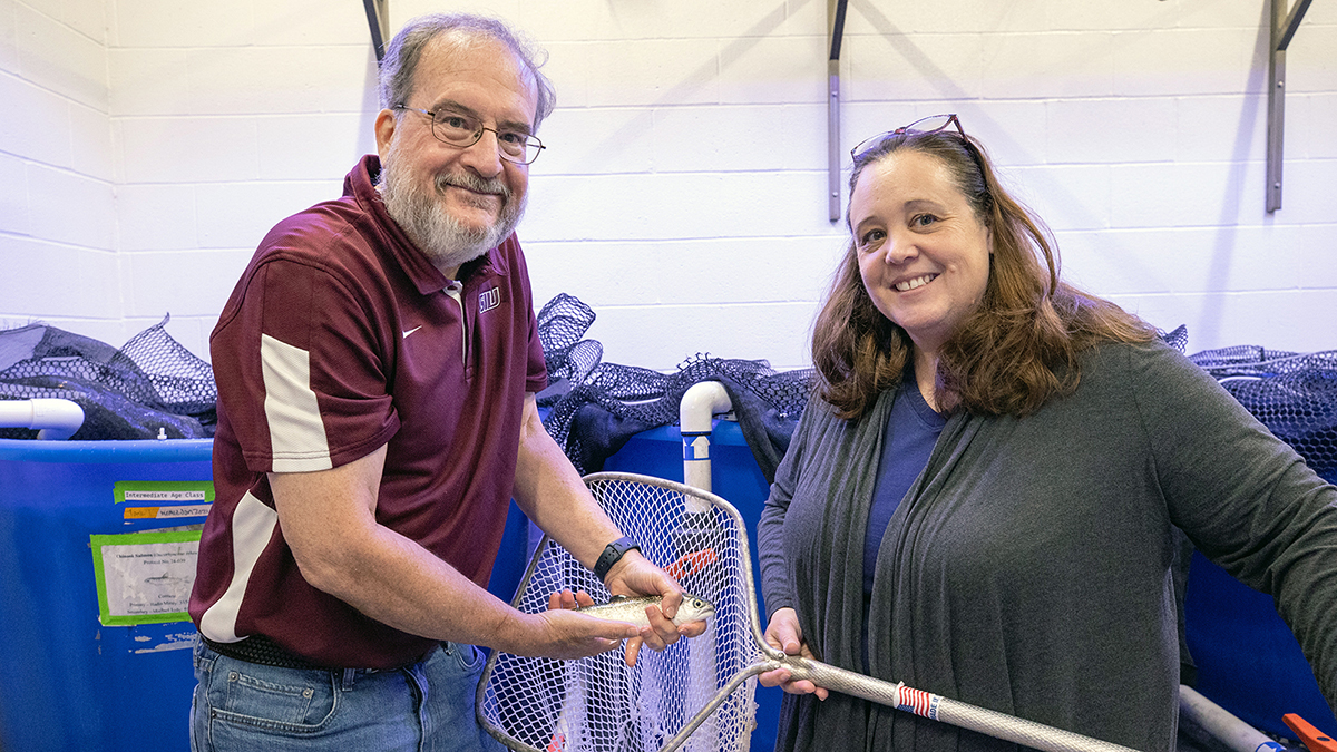 A man and woman stand side-by-side while one holds a small salmon.