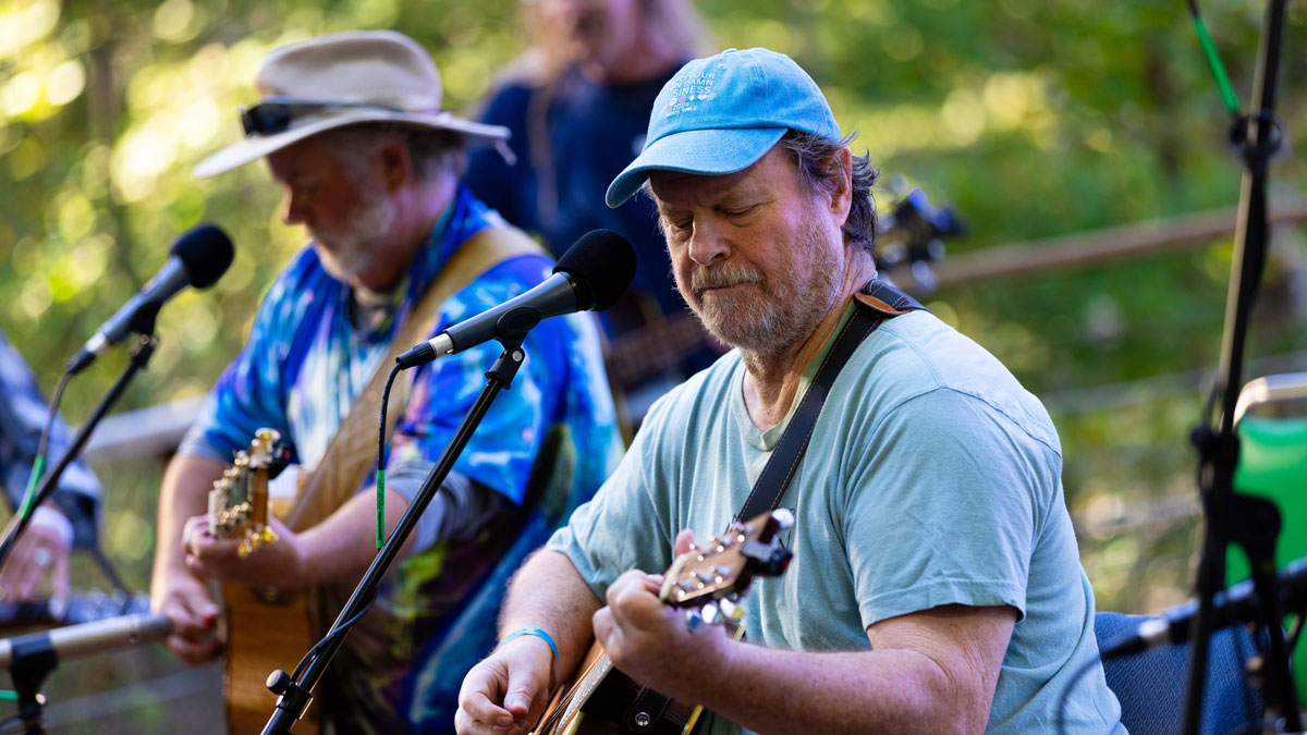 Two men play guitars at an outdoor concert. Another musician is visible in the background. They are surrounded by green foliage.
