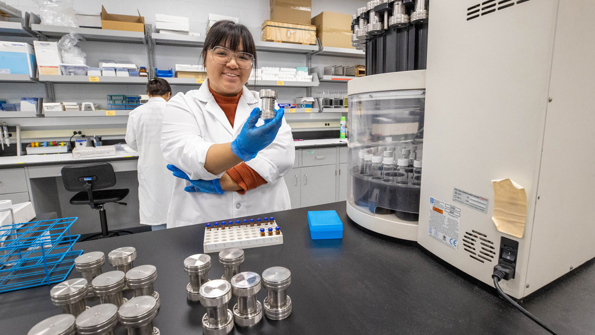 Cristina La is in a lab. She is holding a sample in a small silver container. There are more silver containers in front of her and a colleague is seen in the background.