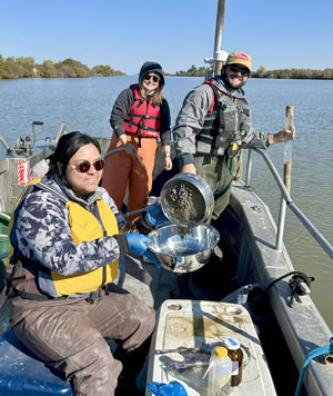 Three people are on a boat conducting research. Cristina La is pouring collected sediment into a bowl.