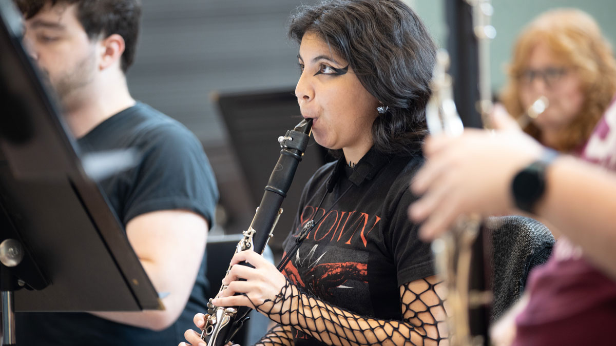 A woman, dressed in a black shirt and bold black eyeliner, plays a clarinet. Other musicians are seen around her.