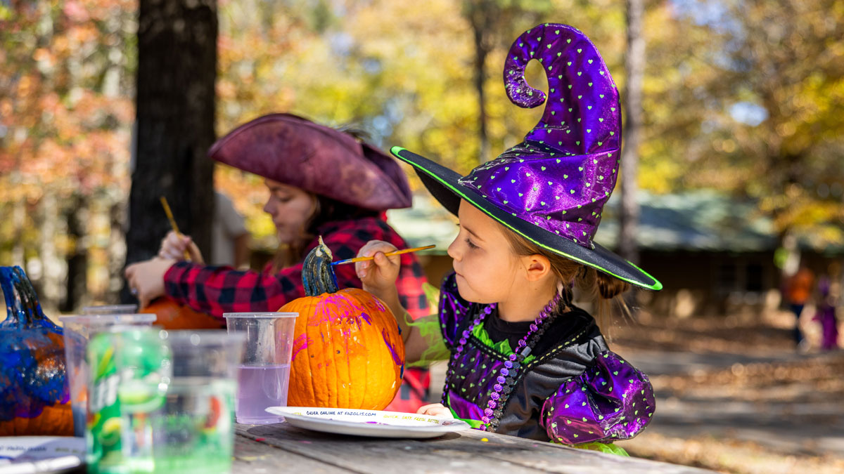A little girl wearing a witches costume is painting a pumpkin.