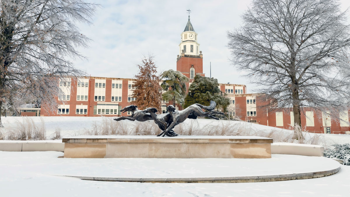 A snowy landscape at SIU Carbondale. A statue of 3 running salukis, at Alumni Plaza, is visible with Pulliam Hall in the background.
