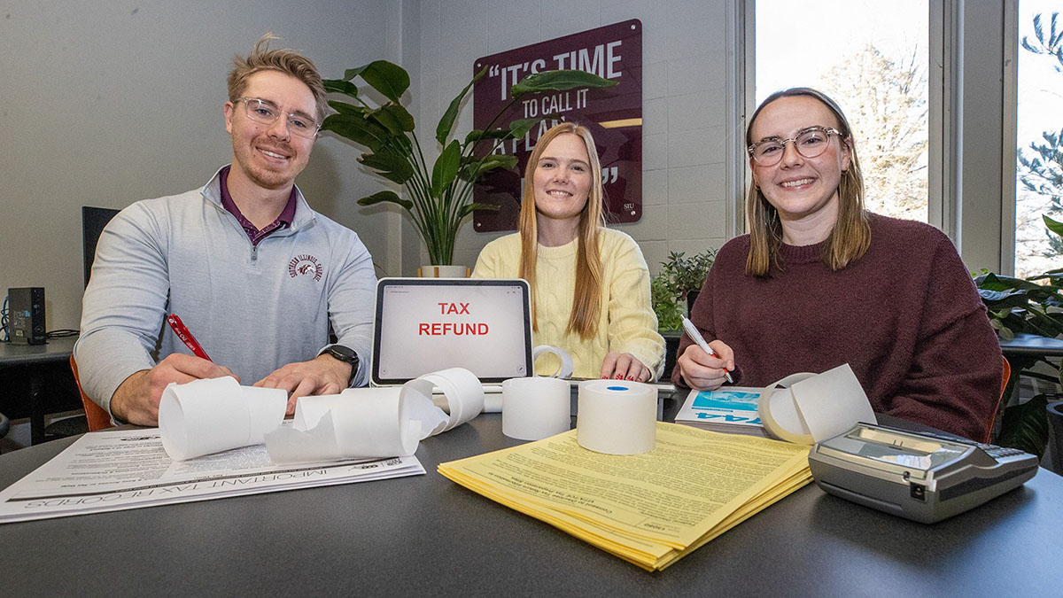 VITA tax preparers are seated at a table covered with papers, receipts, a tablet, and a calculator. The tablet screen says Tax Refund.