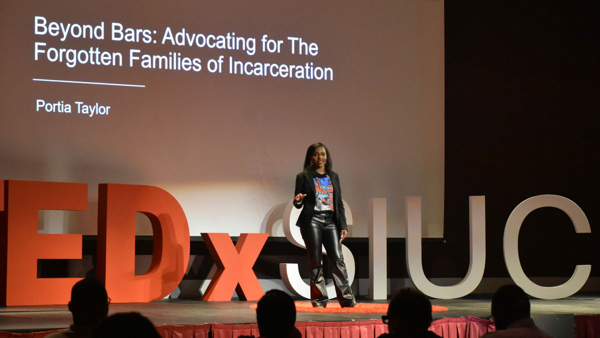 A woman is on a stage. Behind her it reads Beyond Bars: Advocating for The Forgotten Families of Incarceration. By Portia Taylor.