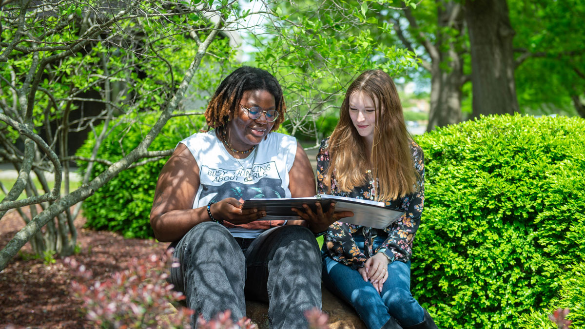 Two women look at a book while seated outside.