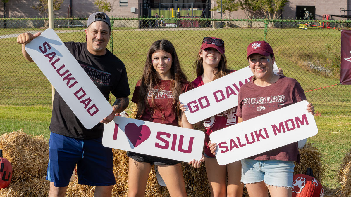 A Saluki family celebrates on Family Weekend at SIU Carbondale. They are holding signs that say Saluki Dad, Go Dawgs, and Saluki Mom.