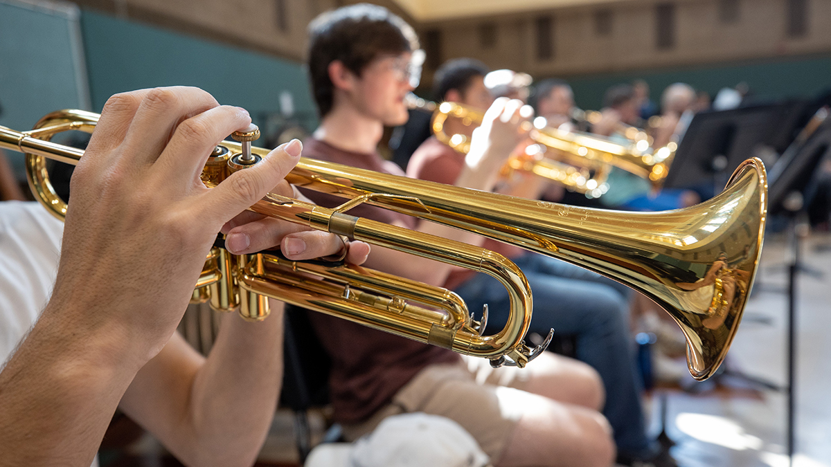 Close up image of a person playing a trumpet. A row of trumpet players are visible in the background.