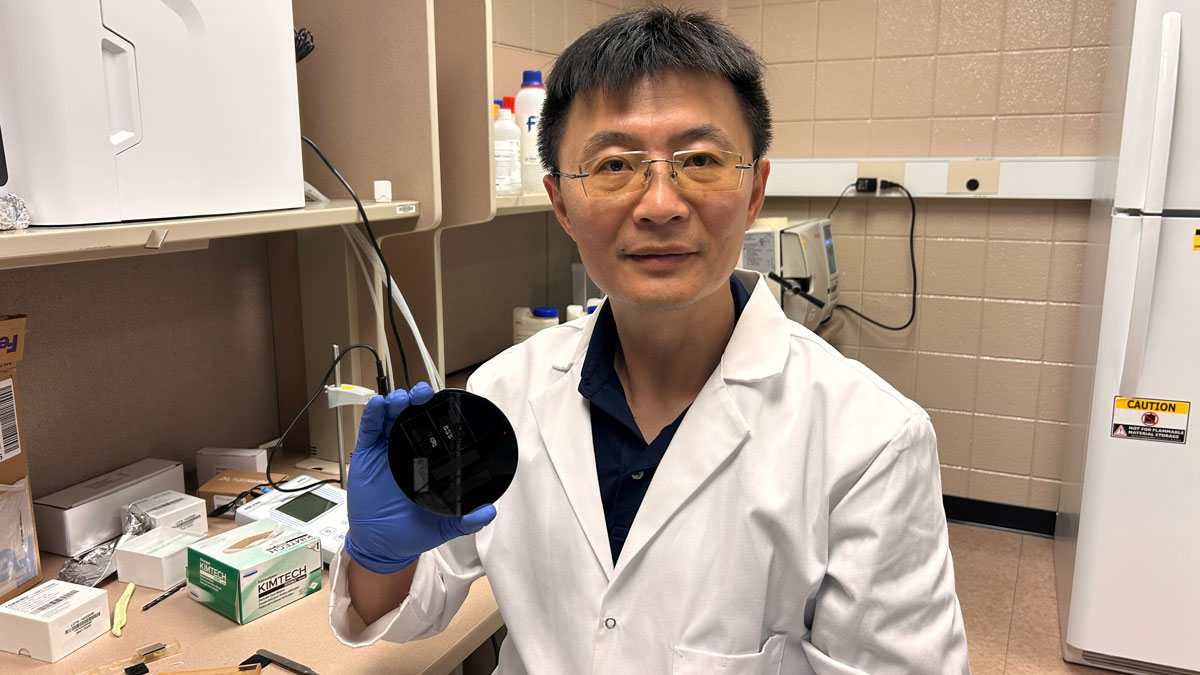 A professor holding a microfluid chip. He is wearing glasses, a white lab coat and blue disposable gloves.
