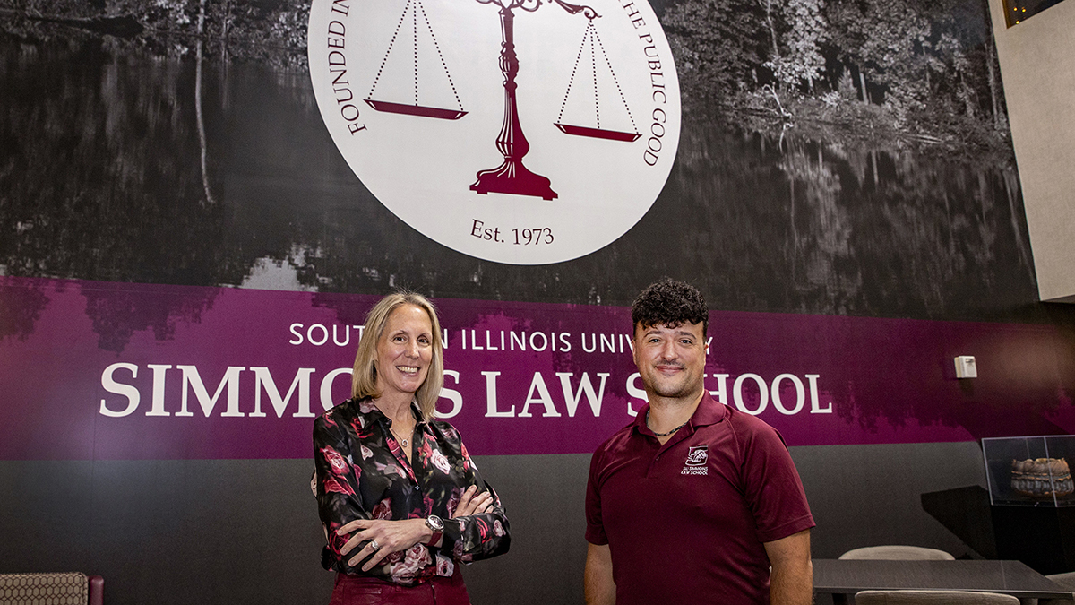A smiling woman with folded arms and a man in a maroon polo shirt stand together in front of a Southern Illinois University Simmons Law School wall display.