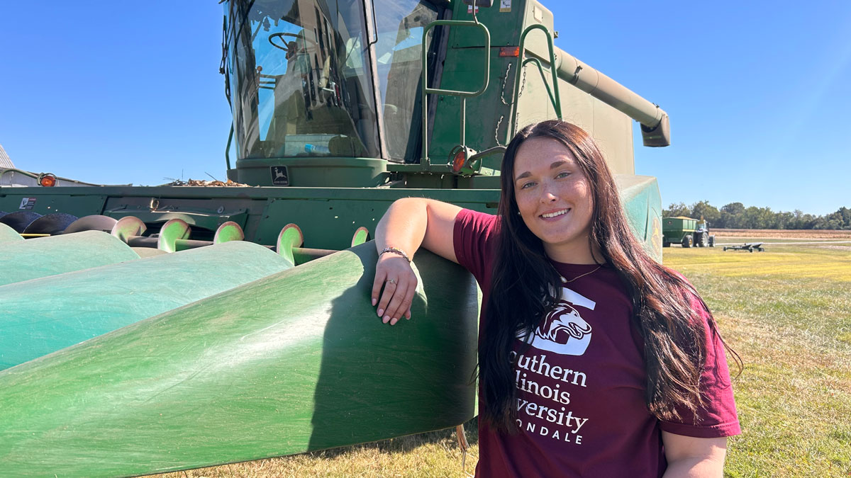 Hannah Humphreys stands next to a combine.  Her maroon shirt reads, Southern Illinois University Carbondale.