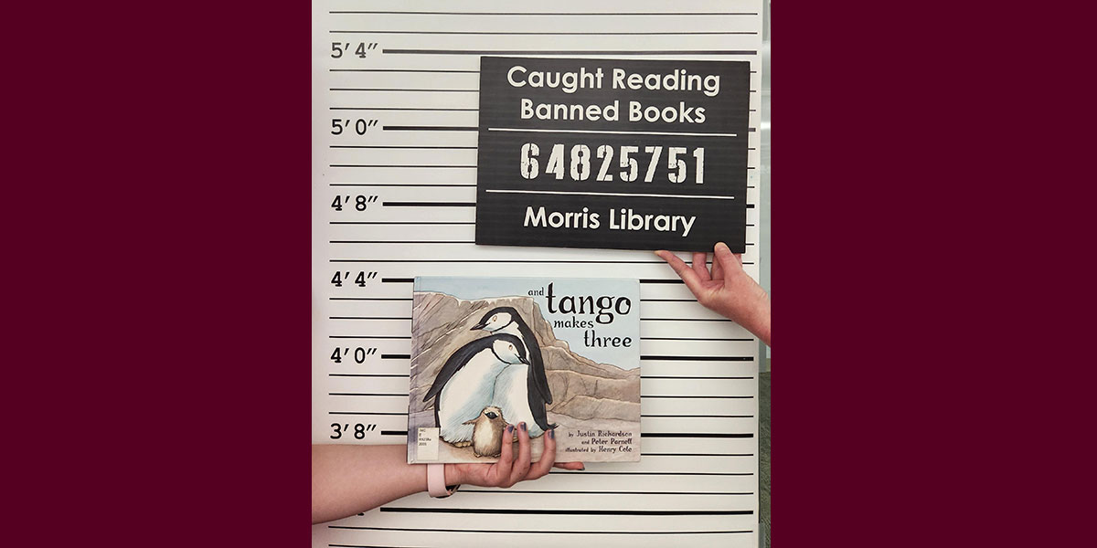 A close-up photograph features a person holding the children's book And Tango Makes Three against a background designed to look like a police mugshot lineup wall. Above the book is a black sign that reads Caught Reading Banned Books, followed by the identification number 64825751, and Morris Library at the bottom.